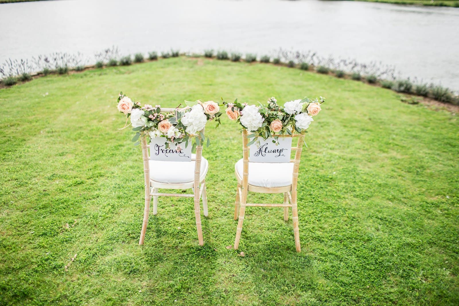 Two ceremony chairs adorned with roses and greenery beside a calm lake